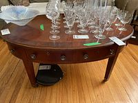 Front and top view of an oval coffee table with glassware on top, showing the inlay wood top and single drawer with metal pulls.