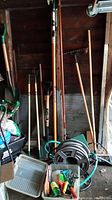 Overview of gardening tools standing against a wooden wall including rakes, shovel, broom, and hose reel with green hose coiled.