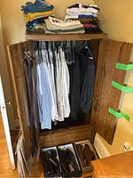 Opened wooden wardrobe with hanging dress shirts, suits, pants in dark colors, belts hanging on door, shoe boxes with black and brown dress shoes below
