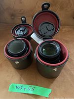 Two Nikkor lenses seated in red velour–lined black cylindrical cases with a green tape note reading “105 + 55.”
