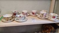 Seven vintage teacups with matching saucers arranged on a shelf, showcasing various floral designs and gold accents.
