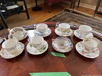 Eight teacups and saucers displayed on table, various floral patterns and brands visible, some with gold trim.