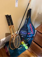 Photo showing two vintage wooden Slazenger tennis rackets leaning against the wall with Dunlop tennis and squash rackets in covers, a golf driver, multicolored tennis balls, goggles, and a green sports bag on the floor.