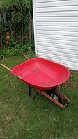 Red metal wheelbarrow set on grass near white building and garden. Shows wheel, wooden handles, and large basin.