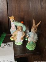 Four Beatrix Potter character figurines displayed on dark wooden shelf. From left to right: a fox in a pink jacket, Jemima Puddle-Duck in blue bonnet, small brown animal character, Peter Rabbit in blue jacket.
