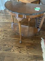 Photo showing side view of vintage demilune table with visible wear marks on top and lower shelf, carved floral motifs on leg apron, hardwood floor background.