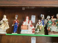 Five distinct ceramic figurines displayed on a wooden shelf, including a lady in a flowing dress, a soldier, dogs on a barrel, and two other figures.