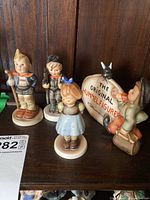 Photo showing four Goebel Hummel figurines placed on a dark wooden surface. Three are children figures; one girl dressed in blue with a bow, two boys wearing traditional clothing, and a decorative sign figurine.
