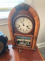 Full view of the wooden mantle clock showing the clock face, wooden frame, painted glass panel, and winding key in front.