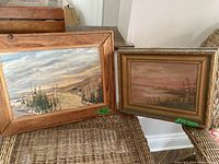 Two framed landscape paintings displayed side by side on a woven chair; the larger painting on the left shows a cloudy sky with evergreen trees and hills; the smaller painting on the right shows a riverside landscape with pink sky