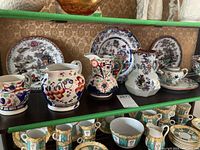 Shelf showing multiple hand painted Asian and English pottery items including pitchers, cups, and plates with floral and Asian landscape motifs.