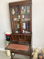 Front view of wooden secretary desk with glass display cabinet, showing fold-down writing surface with inset padded section and visible wear.