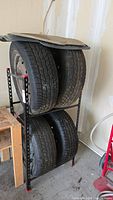 Four tires on rims stacked on a black metal rack in a garage, with floor mats resting on top.