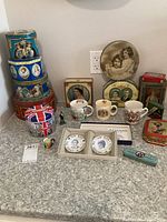 Full display of Royal Family memorabilia and English decorative tins arranged on countertop including mugs, plates, tins, and toothpick holder.