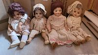 Five vintage dolls placed sitting against a cardboard box on a cement floor. Dolls show signs of wear and damage.