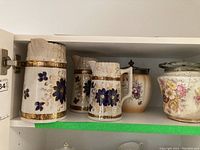 Three vintage cream-colored ceramic pitchers with blue floral and gold detailing displayed on a shelf alongside two lidded bowls/jars with floral patterns.