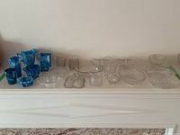 Overview of 21 antique Sowerby glass pieces on white mantel including blue swirling glass items and clear pressed glass items.