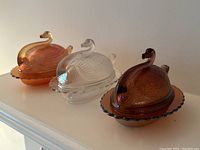 Front angled view of the three swan shaped lidded glass dishes showing orange carnival glass, clear glass, and amber carnival glass finishes.