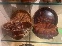 Four large pieces of pottery displayed on a glass shelf: two spatterware bowls, one spatterware plate, and one brown ceramic mold with shell decoration.