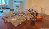 Wide shot of the entire collection showing multiple pressed glass bowls, baskets and vases on wooden table with ambient light from window.