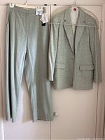 Full view of the suit jacket and pants hanging on white hangers against a white closet door showing the pale green wool fabric and tags attached to pants.