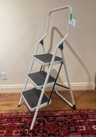 Full side angle view of the 3-step ladder showing white metal frame, safety handle, black non-slip treads on steps, and rubber feet caps.