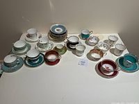 Wide view of all teacups and saucers arranged on a white surface, showing various patterns and colors including turquoise, red, white with floral and gold accents.