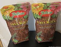 Two sealed bags of Hoffman volcanic lava rock decorative soil cover, showing the colorful packaging with floral graphics and clear product visibility.