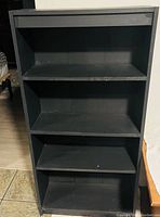 Photo of a black wooden bookshelf with three shelves. The bookshelf stands on a floor, has a simple rectangular design with some wear.
