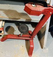 Red metal can crusher with a pedal and spring mechanism, placed on a dusty metal shelf.