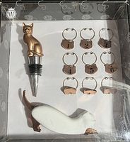 Photo shows a packaged set containing a bronze cat wine bottle stopper, nine small metal wine glass charms with numbers and paw prints, and a white cat-shaped keychain with bronze details.