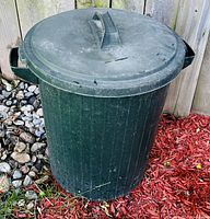 Photo of a green round plastic garbage can with lid placed outdoors on grass and red mulch with rocks and a wooden fence in the background.