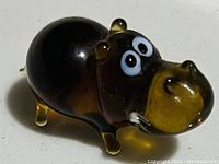 Close-up photo of a small glass hippo figurine showing brown and amber coloration, large white and black eyes, rounded body, ears, and tail.
