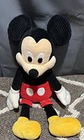 Photo of Mickey Mouse Scentsy plush sitting upright on a patterned carpet with a dark background showing the front details including ears, face, red shorts, and yellow shoes.