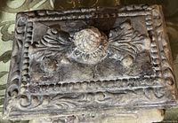 Close-up of the trinket box lid showing ornate floral and scroll carved details in weathered dark material with a rounded central knob.