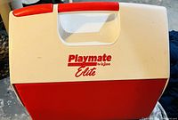 Photo of Playmate Elite camping cooler showing the white top with red handle and red bottom half, branded 'Playmate by Igloo Elite' on front.