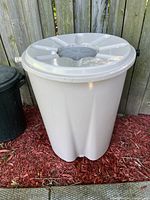 Large gray plastic rain barrel with a gray lid that has a circular opening in the center, pictured outdoors on red mulch against a wooden fence.