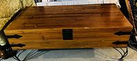 Closed solid wood blanket box on metal scroll legs, showing wood grain and black metal hinges and lock plate on front.