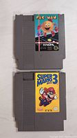 Two gray Nintendo NES cartridges stacked vertically showing front labels: Pac-Man by Tengen on top and Super Mario Bros 3 below.