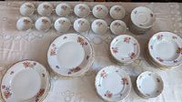 Wide view of 12 sets showing dinner plates, salad plates, bowls, cups, and saucers arranged on a white tablecloth. Each item features rose floral prints and gold rims.