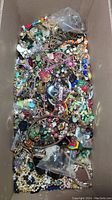 Overhead view of box full of assorted costume jewelry items including beads, bracelets, necklaces, pins, and earrings in various colors and materials.