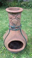 Front view of the chimnea showing the wide mouth opening and grapevine design on the neck and body, placed on grass.