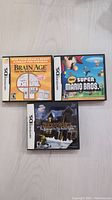Front view of three Nintendo DS game cases: Brain Age, New Super Mario Bros., Nancy Drew: The Hidden Staircase laid on light wood floor.