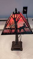 Front side of the lamp lit up showing moose and tree silhouettes on stained glass shade with a mix of orange, pink, and purple colors.