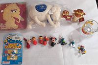 Photo showing the Sesame Street Big Bird needlepoint kit, Serta white sheep plush in plastic, Garfield ornaments and glass mug, along with Smurfs and Fisher Price Little People figures spread out on a white surface.