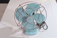 Front view of teal Torcan fan showing blades, wire guard, and base.