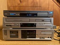Image showing stacked Samsung P231 DVD player (top), Electrohome DVD/VCR player (middle), and Akai HX-A101 cassette player (bottom) on wooden floor.