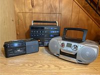Photo of three portable stereo cassette players including a Citizen AM/FM radio cassette recorder, a black multi-cassette player, and a silver Panasonic boombox on wood floor against wood wall.