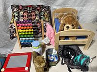 Full view of the toy lot showing abacus, wooden bed with teddy bear, red magnetic drawing board, stuffed animals, headphones, decorative pillow, and other assorted items.