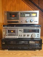 Four stacked electronic units: Fisher black dual cassette player at bottom, Technics cassette player above it, Toshiba DVD player in slim black design, and Akai cassette player on top with silver front and analog meters.